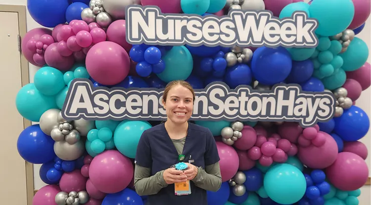 a person holding a cupcake standing in front of a huge ballon display with a sign reading "Nurses Week Ascension Seton Hays"