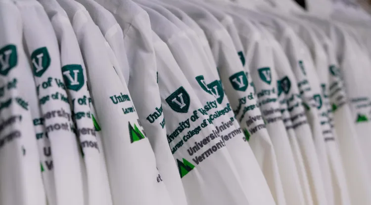 a rack of hanging white medical coats with the UVM Larner College of Medicine and the UVM Health insignia embroidered on the upper shoulder