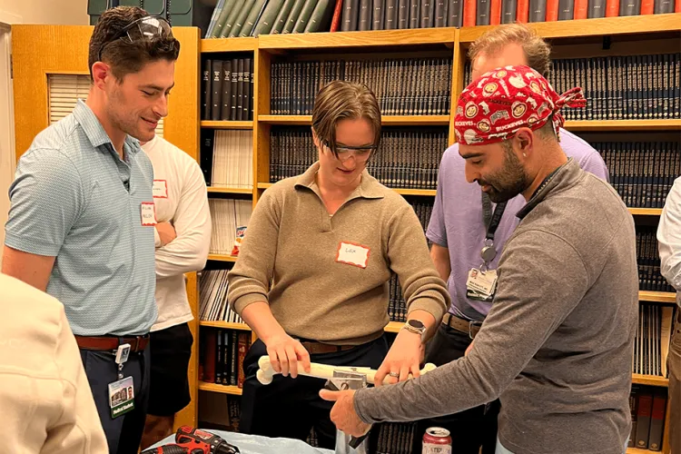 Several people gather in a room with bookshelves in the background all looking at a fibula bone model. 