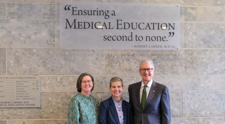 3 people standing in front of a sign mounted on a stone wall that says, "Ensuring a medical education second to none."
