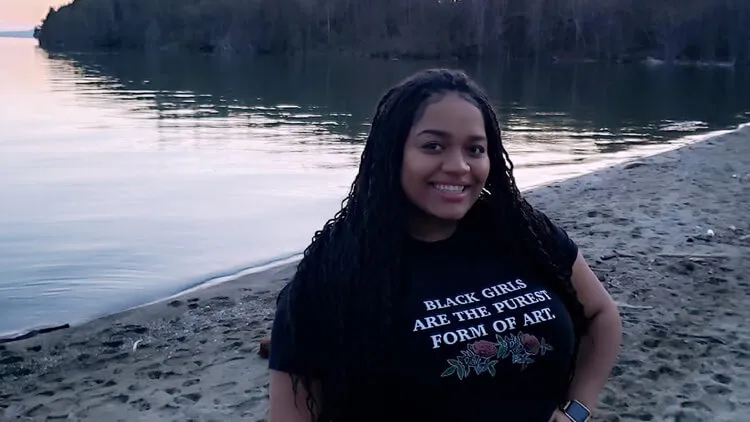 Person standing on a sandy beach beside a lake with trees in the background.