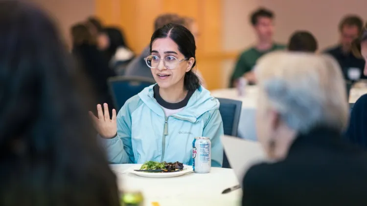A person in a blue jacket and glasses shares thoughts at a table while others listen