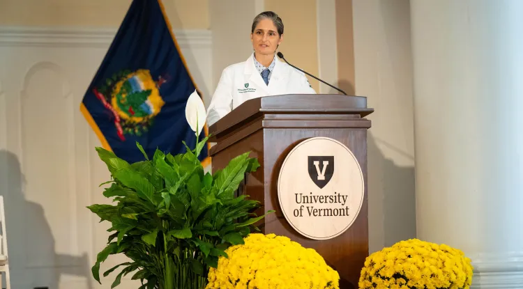 a person in a white coat at a podium with a seal showing a big "V" and the words "University of Vermont", decorated with plants and flowers and a Vermont State flag in the background