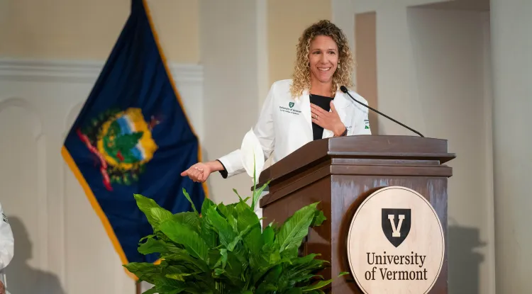 a person in a white coat speaking at a podium with a seal with the letter "V" and the words "University of Vermont", decorated with a plant and the Vermont State flag in the background