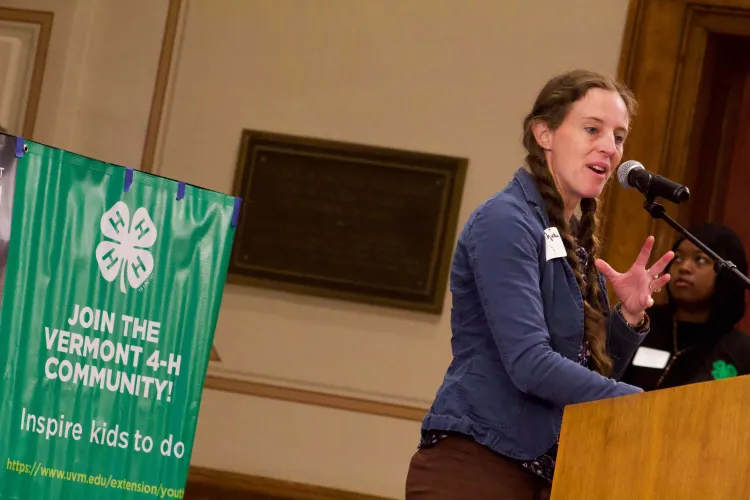 A white woman with long dark blonde braids speaks at a podium with a green Vermont 4-H poster in the background.