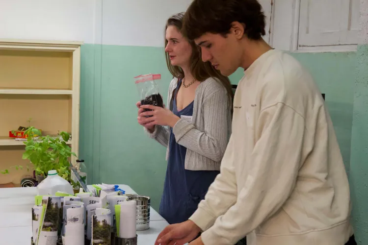 Two college students stand by a table with a bag of soil, potted plants and cans with papers curled up inside them