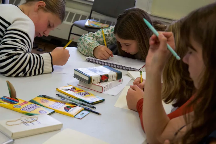 Young people around a table draw the body shape of birds with colored pencils.