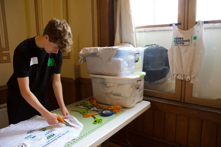 A young man in a black shirt with a green four leaf clover on it, cutting up a t-shirt by a window where another t-shirt hangs and reads: "teens reaching youth."