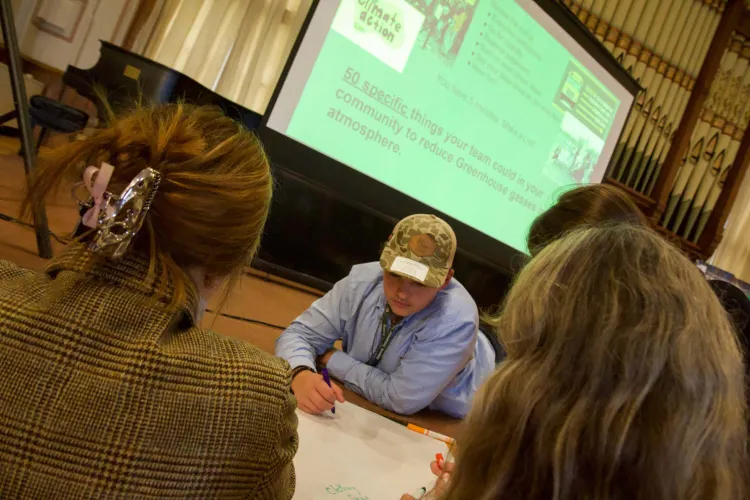 Young people gather around a giant piece of paper underneath a large screen providing instructions to list 50 things students could do to reduce greenhouse gases in their communities.