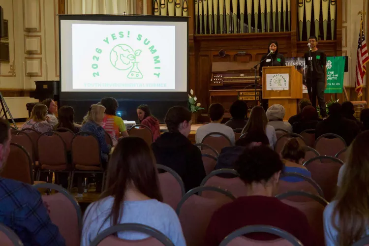 Two teenagers stand at a podium next to a screen reading 2026 YES Summit in front of a room full of people.