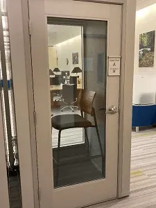 One-person metal pod with glass door and large window, inside a larger room.
