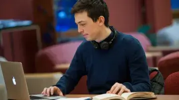 student sits at a desk in a library looking at a laptop