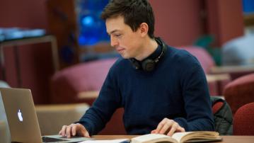 student sits at a desk in a library looking at a laptop