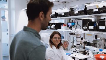 student in a white lab coat talks to a person standing nearby at a lab workbench