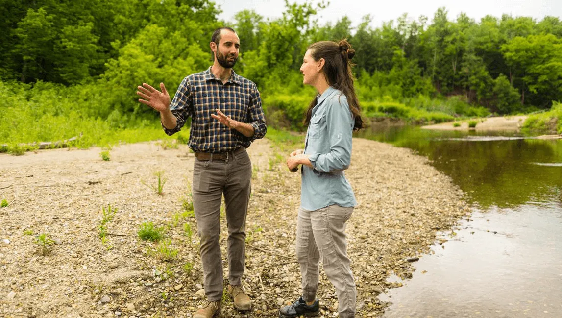 Jesse Gourevitch standing on a river bank talking to a female researcher