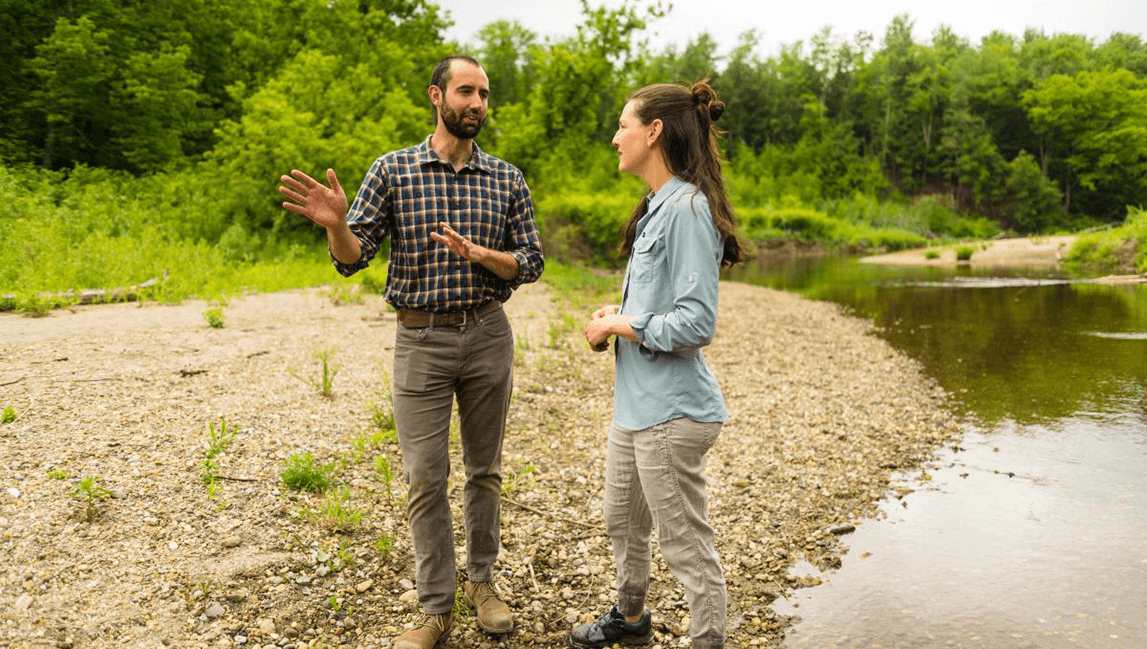 Jesse Gourevitch standing on a river bank talking to a female researcher