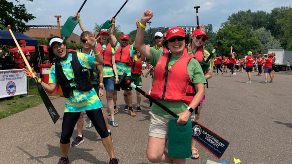 Several people outdoors wearing colorful clothes and life jackets, holding paddles, arms raised, smiling