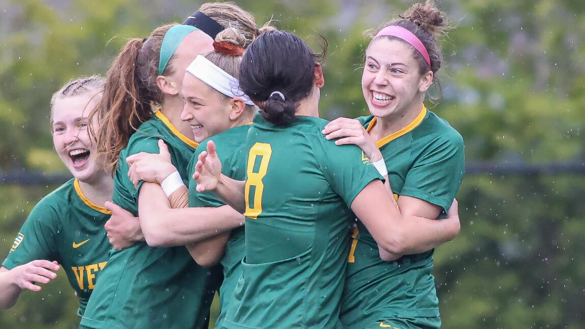 A group of people wearing green and gold soccer uniforms engaged in a group hug while smiling