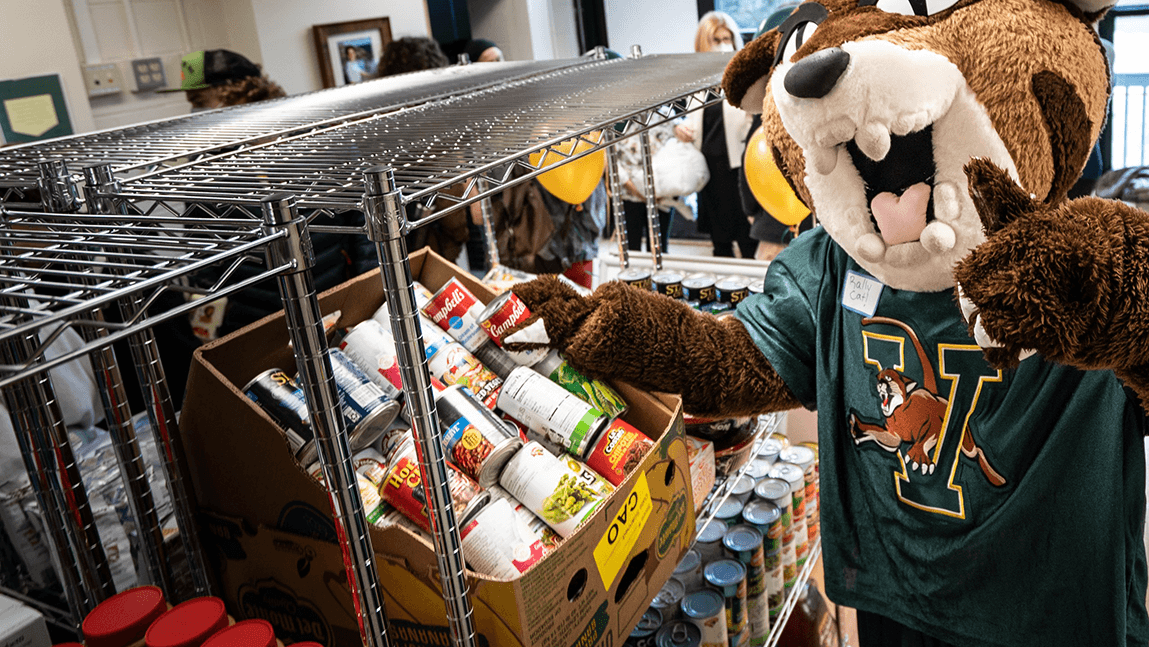 Rally Cat poses for the camera in front of shelves of non-perishable food items in UVM's student food pantry.
