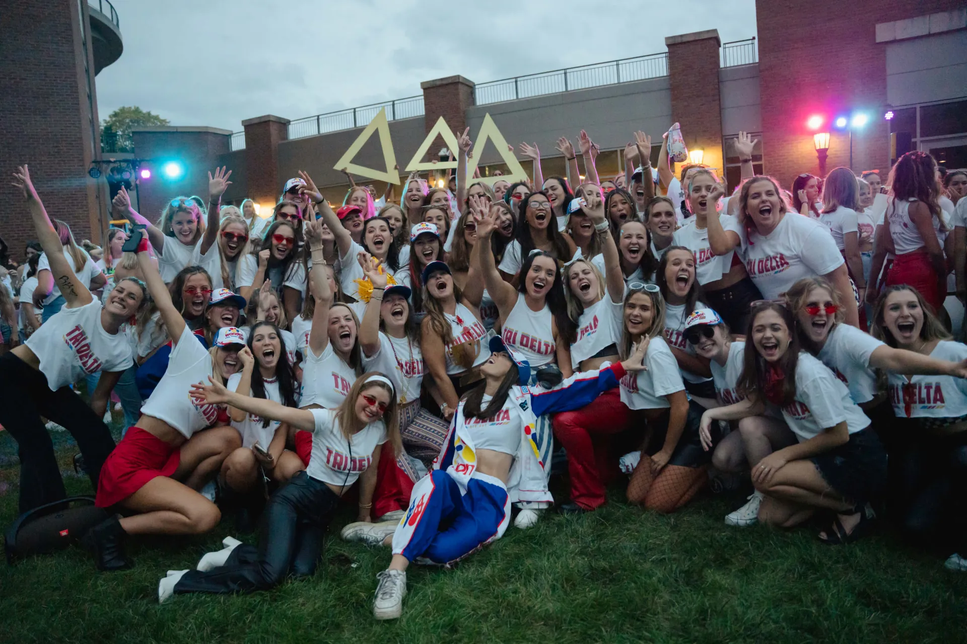 Outside the Davis Center at dusk, a group of more than 50 young women all excitedly pose for the camera, their arms up in celebration, smiles on their faces. They all are wearing the same white t-shirt that reads in big bold lettering "Tri Delta"