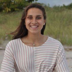 Ashleigh Angle. A person with dark hair wearing a white shirt with pink strpes sits on a beach with grass in the background smiling at the camera. 