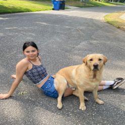 Ella Weigel. A person with dark hair sits wearing a dark top and blue shorts sits on the ground. A yellow labrador retriever sits on their legs. 