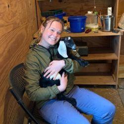Lauren Smathers. A person with light brown hair, wearing a green jacket and jeans, hold a baby goat and smiles at the camera.