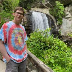 Matt Jacobs. A person with dark hair, in glasses, a tie-dye shirt, and dark shorts stands in front of a waterfall, smiling at the camera