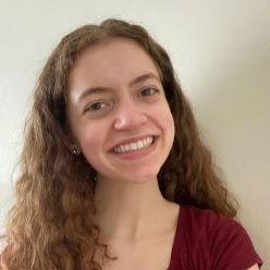 Sophie Unger. A person with dark curly hair and a red shirt looks at the camera and smiles.