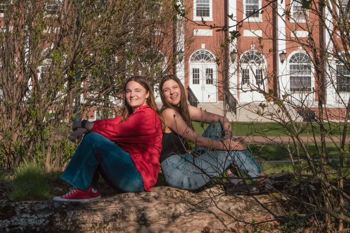 two young woman, one in red jacket and the other in a black tank top sitting on large rock in front of brick building in early spring