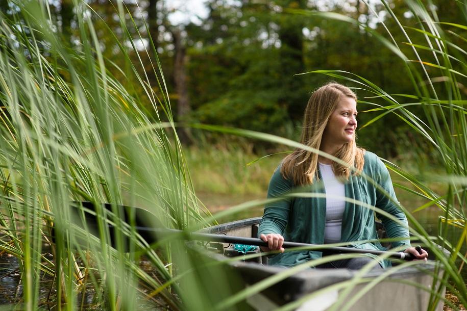 A person is sitting in a canoe holding an oar, surrounded by reeds and other green plants.