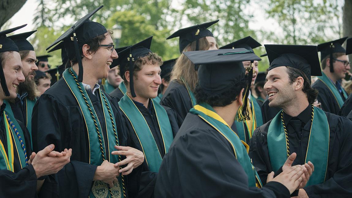 Graduates clap and smile in the shade of trees.