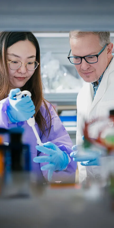 An Asian student conducts an experiment while a faculty member looks on.