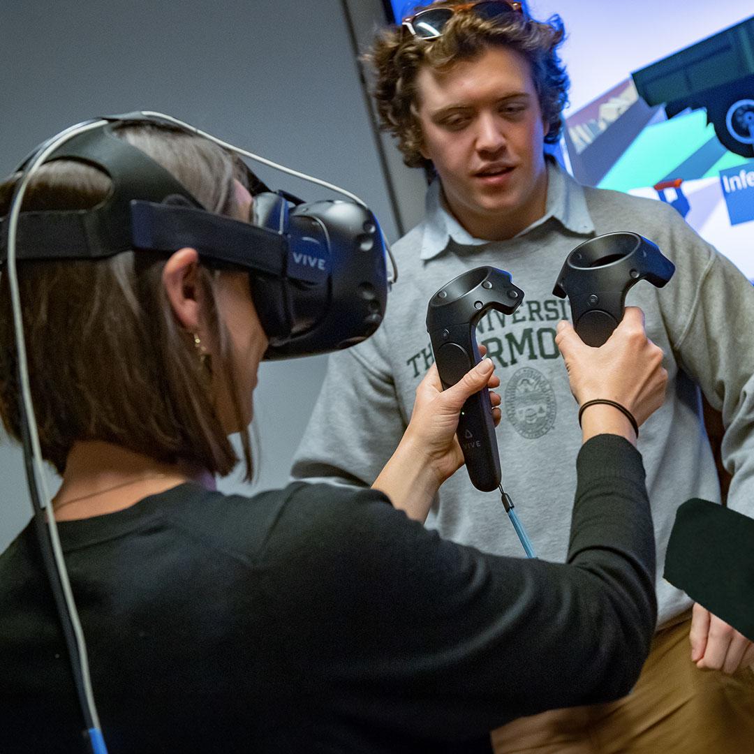 a student wearing a vr headset holds controllers while another student looks on