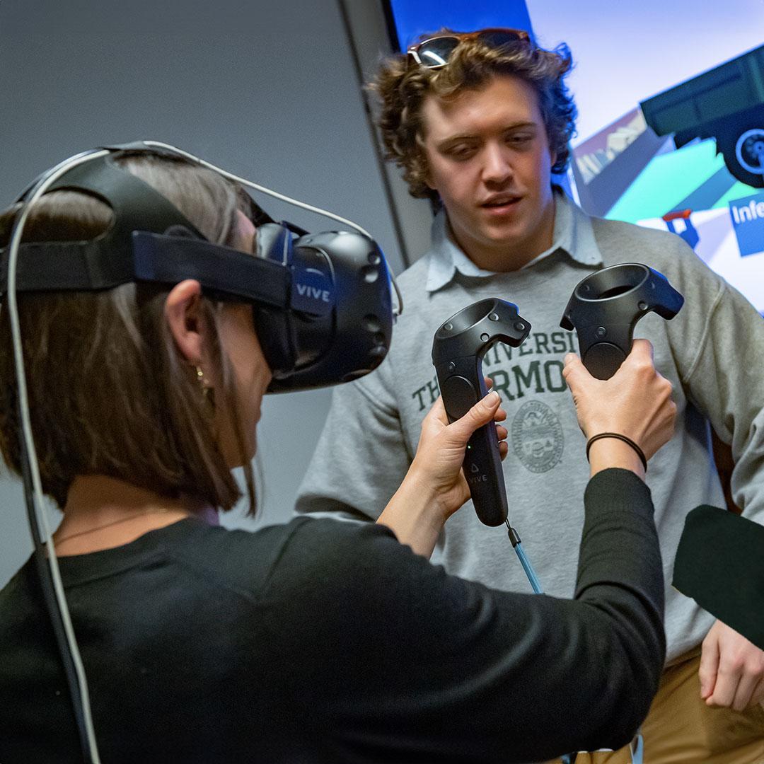 a student wearing a vr headset holds controllers while another student looks on