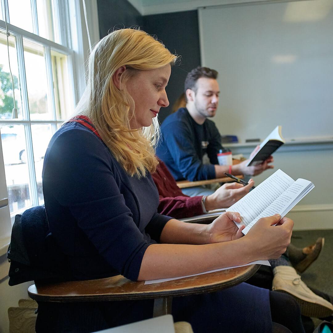 Two people sit in desks holding open books.