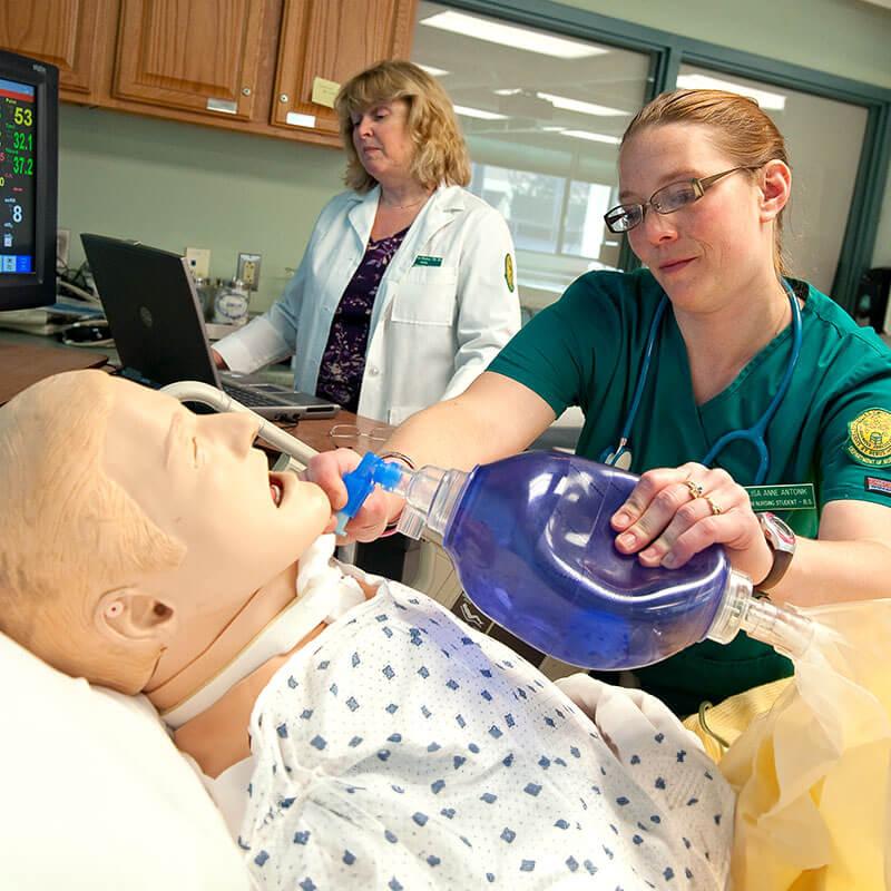 a student works on a test dummy while a professor observes from afar