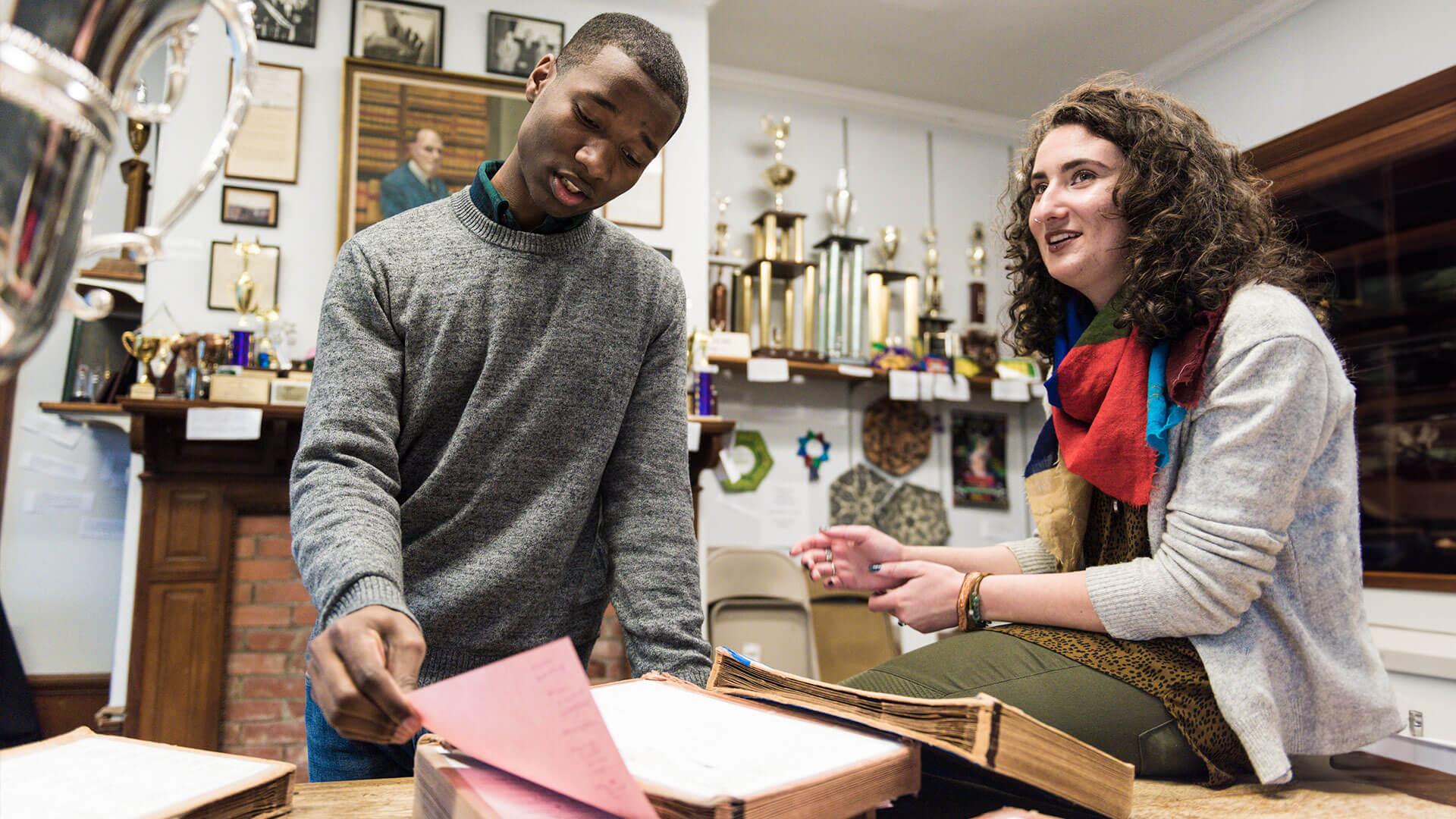 two debate students look through notes in front of a trophy