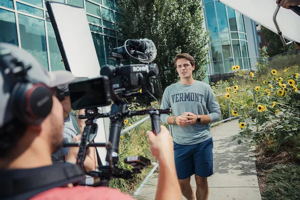 Behind the shoulder of a camera operator filming a student speaking