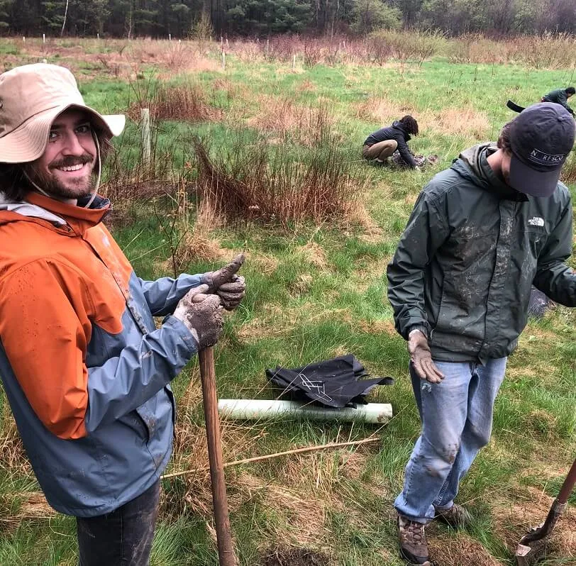 Students on a rainy day digging holes for ecological restoration, one is smiling and giving a thumbs up to the camera