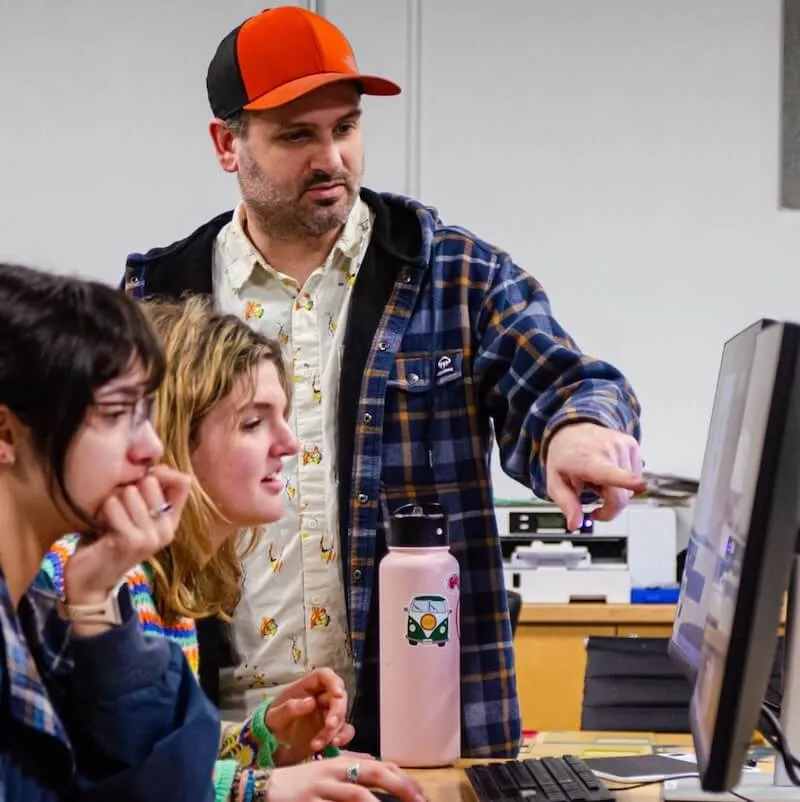 Students in front of a computer are directed by an instructor.
