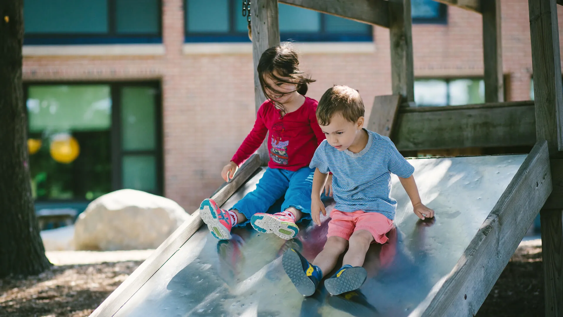 Two children sliding down a slide on a playground outside.