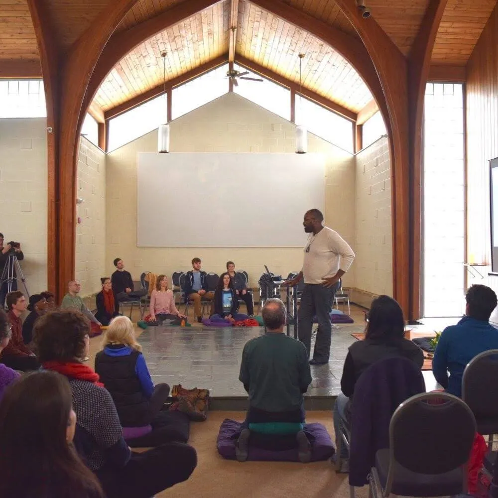 People sit on the floor and in chairs listening to a speaker in the middle of the room on a raised tile floor.