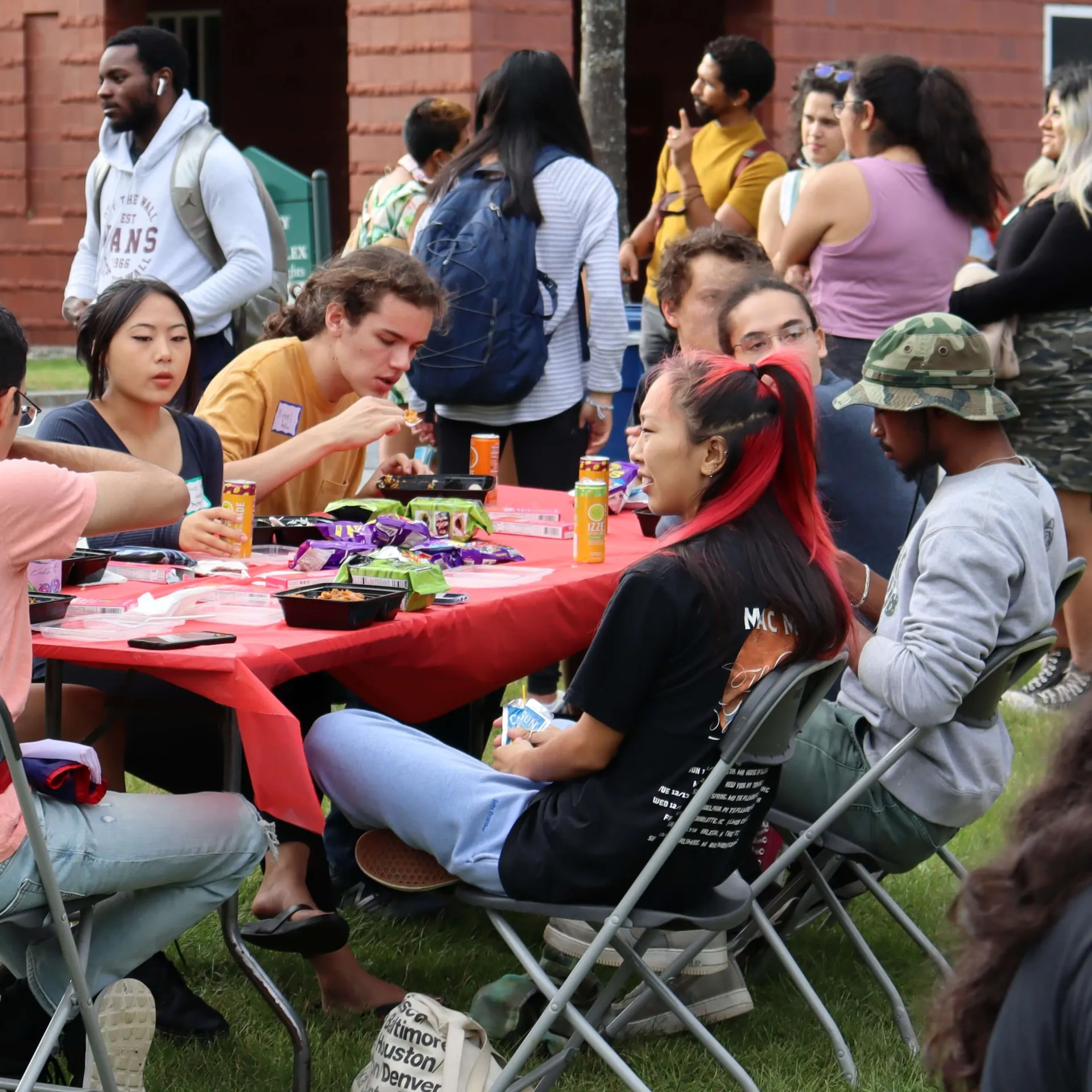 Students of different racial and ethnic backgrounds sitting at an outdoor table eating and talking while more students gather behind them.