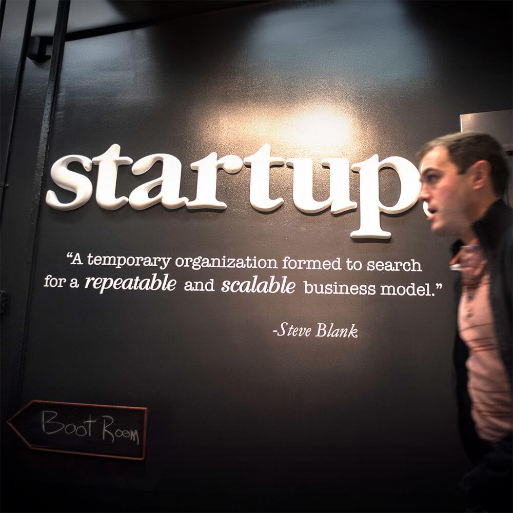 A wall with a sign that says "startups" with a quote by Steve Blank. "A temporary organization formed to search for a repeatable and scalable business model."