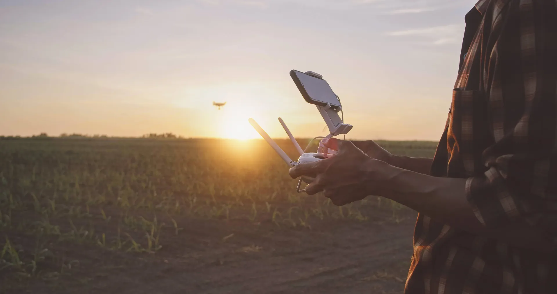 Man in a flannel flies a drone at sunset.
