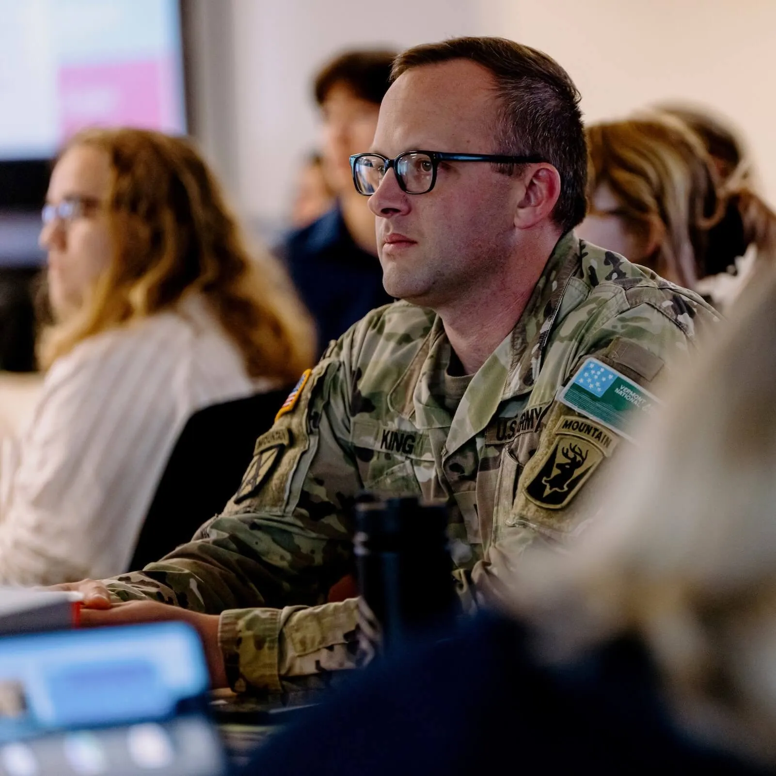 Man in Vermont National Guard uniform sitting at a table, listening attentively to someone off camera