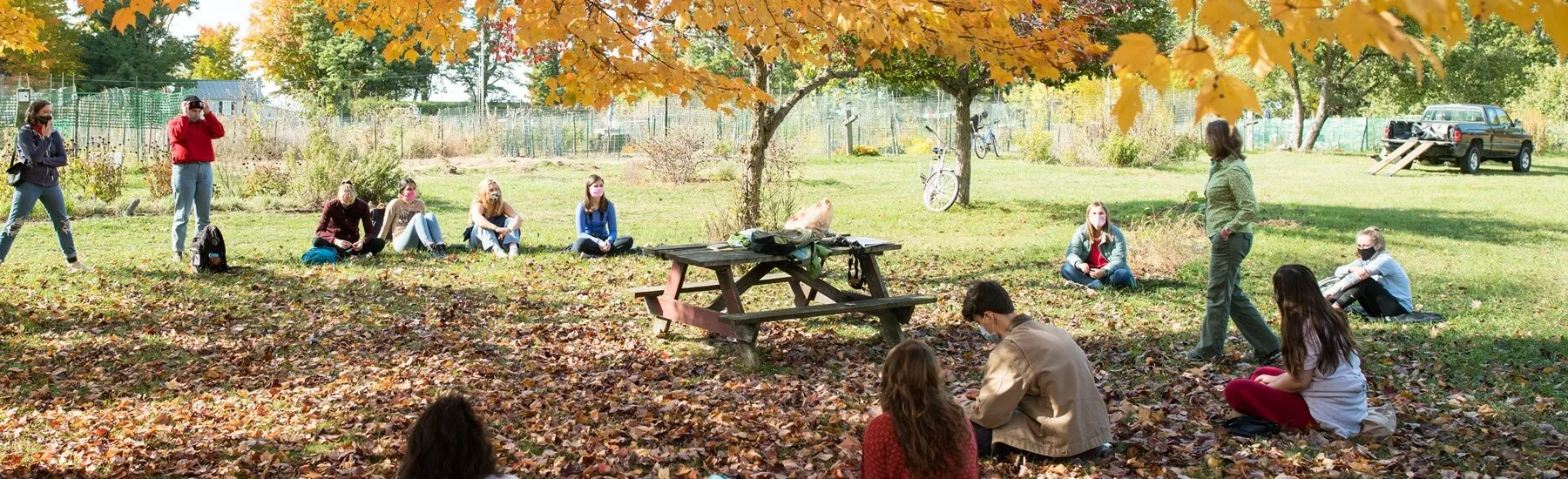 Students sitting outside near a picnic table listening to a faculty member, yellow leaves on the ground and in the tree above them.