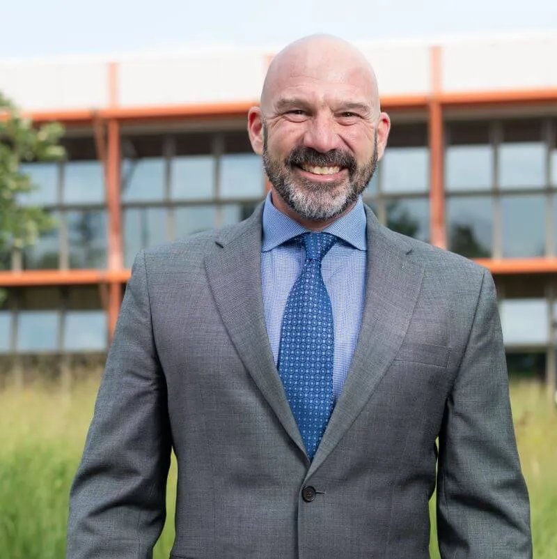 Dean Peter Newman smiling wearing a suit and tie, outside in front of the Aiken Center, the background is blurred