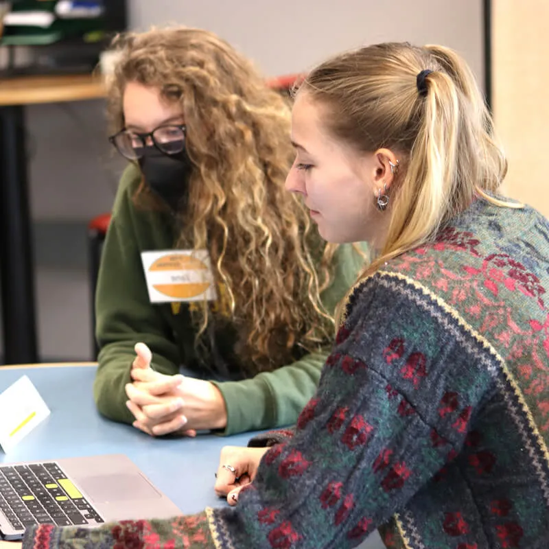 Two women, a writing tutor and a student, sit at a writing center table facing a laptop. One is masked and the other is not.